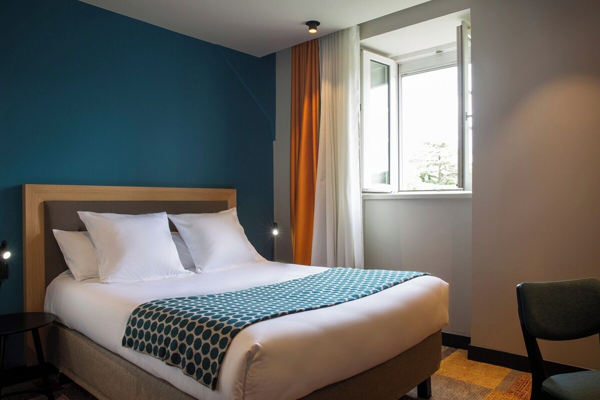 A hotel room with a bed, a chair, a window, and a nightstand.