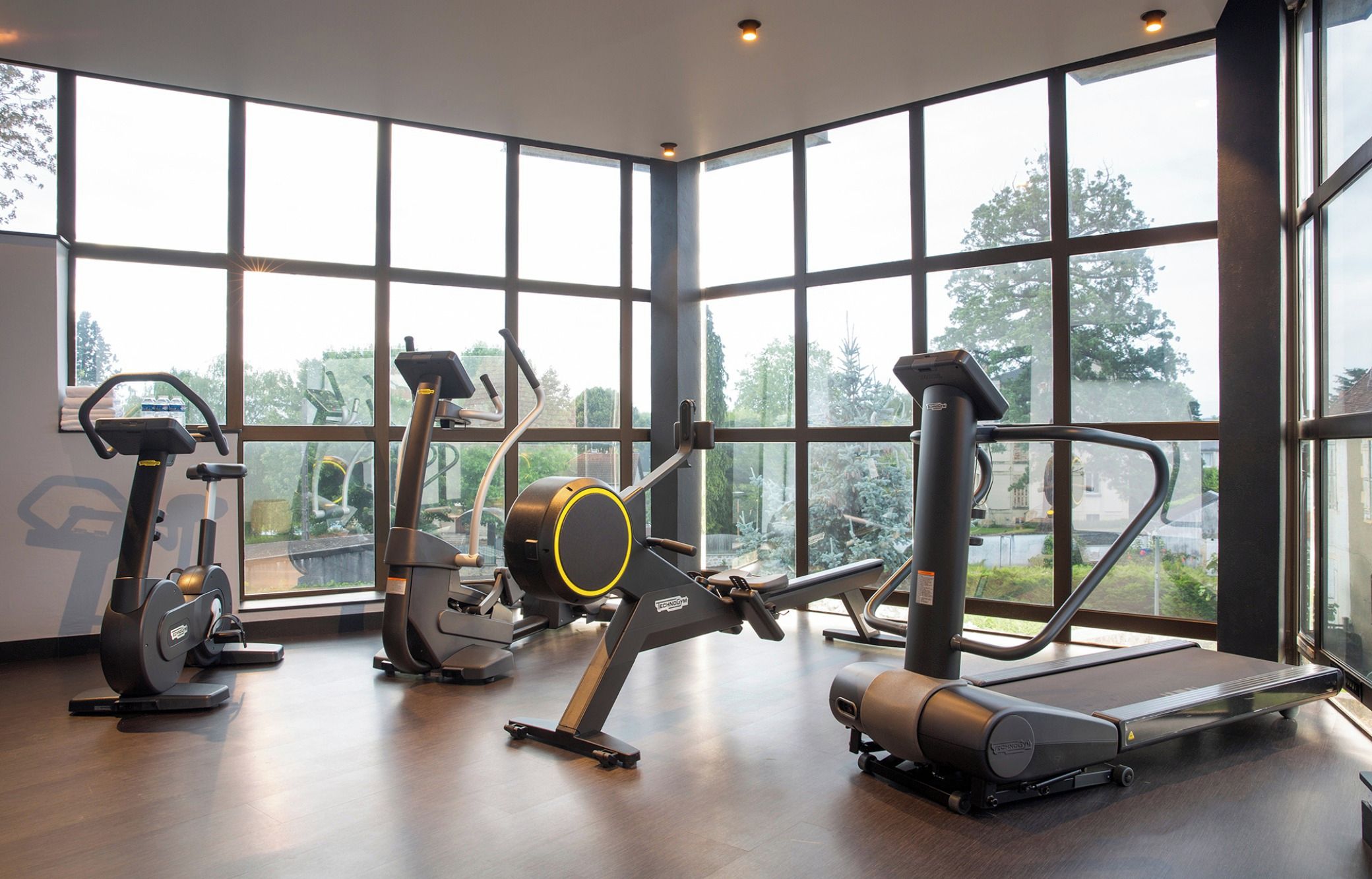 A gym with cardio machines, a rowing machine, and a treadmill, with large windows offering a view of greenery.