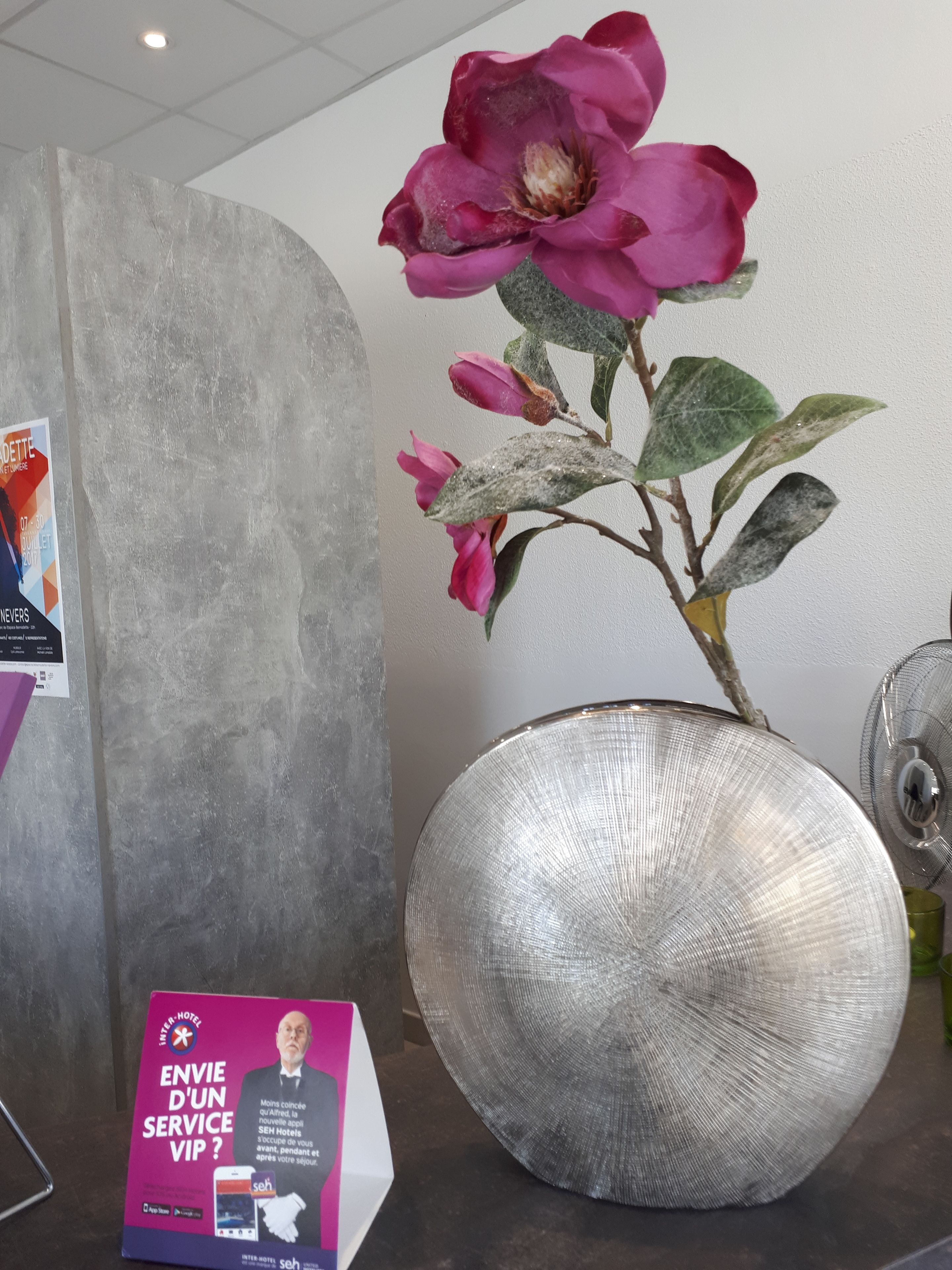 Un grand vase métallique orné d’une composition florale rose, une enseigne publicitaire pour les services de l’hôtel et une vue partielle d’un mur.
