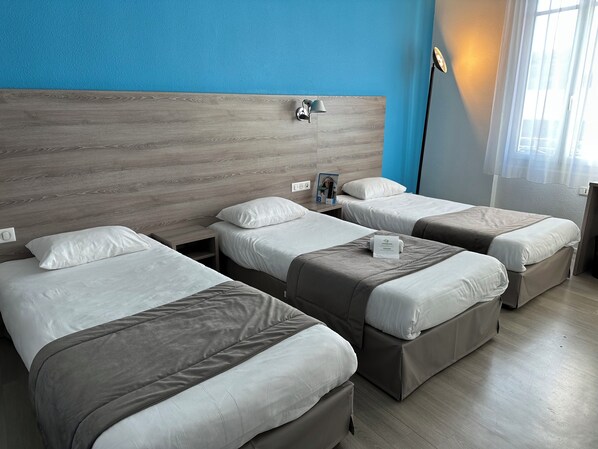 A hotel room with three beds, a wooden headboard, a nightstand with a lamp, and a window with curtains. - BRIT HOTEL CONFORT NEVERS Centre Gare (Nevers)