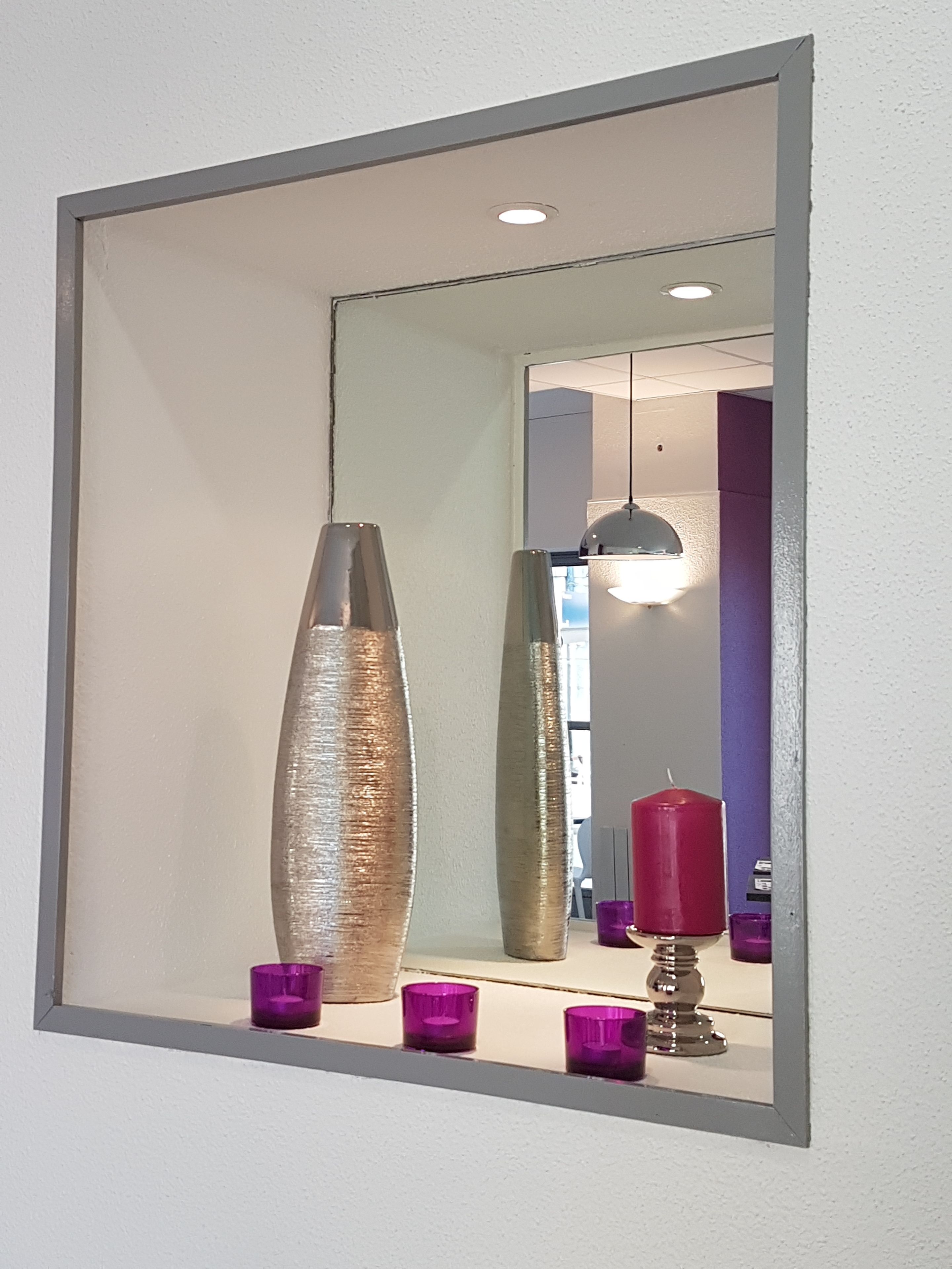 Un miroir doté d’un cadre métallique qui reflète une pièce dans laquelle on aperçoit un grand vase à motifs, une bougie rouge et plusieurs chandeliers violets.