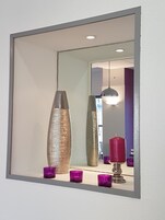 Espejo con marco metálico que refleja una habitación con un jarrón alto y texturizado, una vela roja y varios candelabros morados.