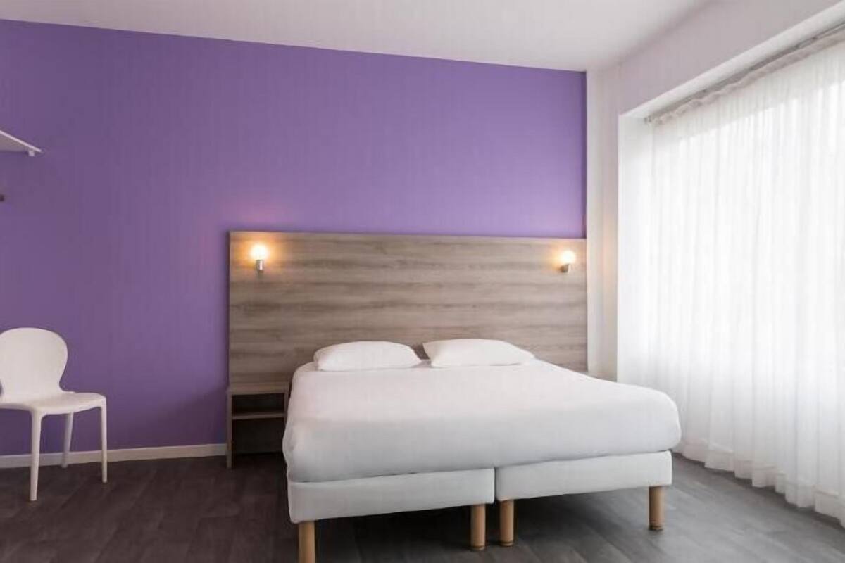 A hotel room with a bed, two wall-mounted lights, a chair, and a window with curtains.