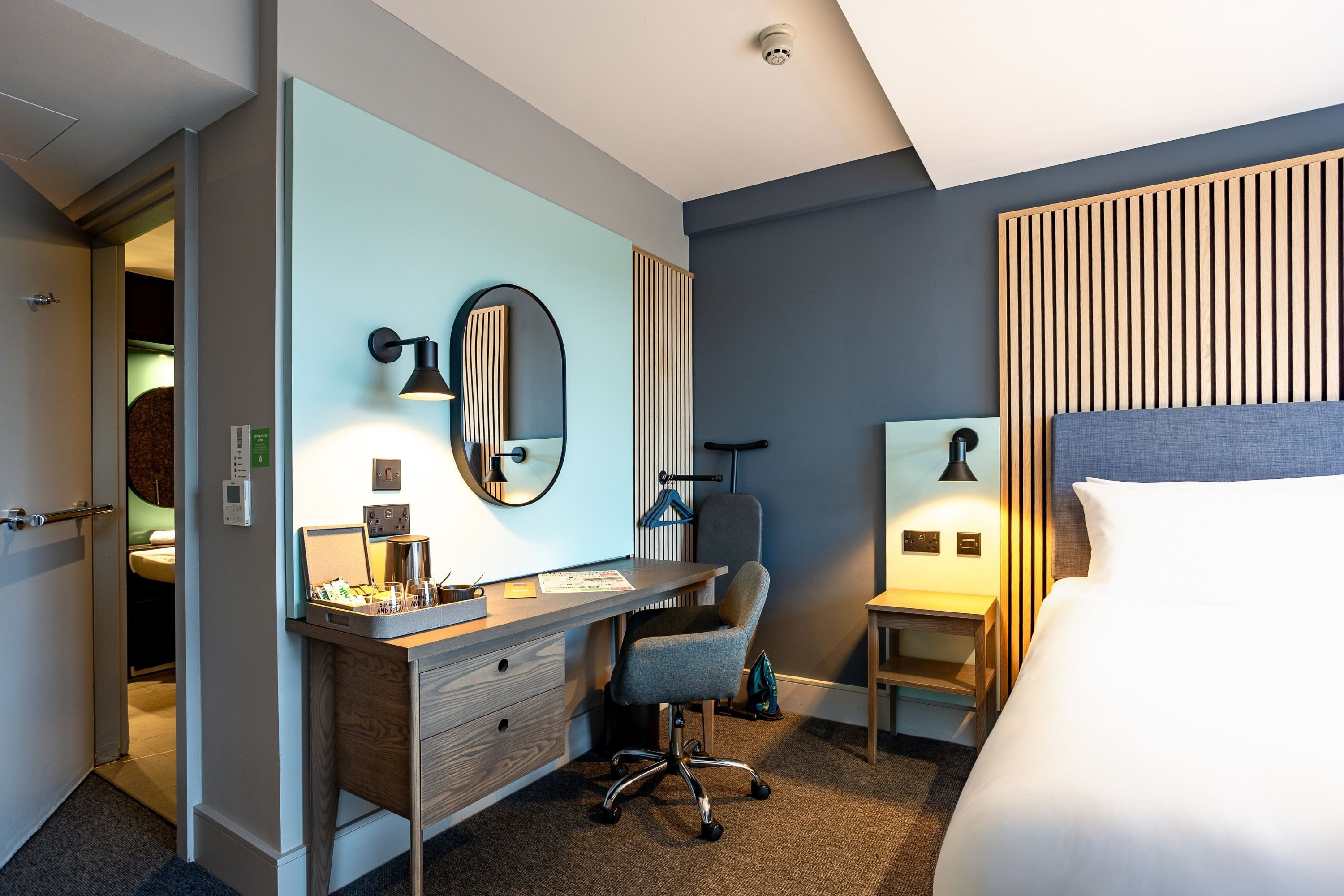 A hotel room with a bed, a desk with a chair, a mirror, and a bedside table.