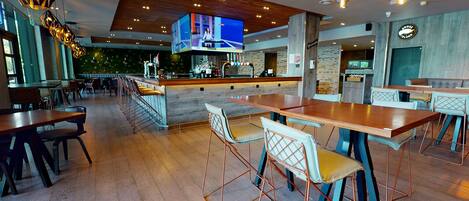 Interior moderno de restaurante con mesas de madera, barra y una gran pantalla de televisión.