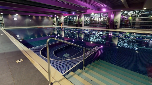 An indoor swimming pool with a blue tiled floor, a metal ladder, and a view of trees through large windows. - Village Hotel Leeds South (Leeds)