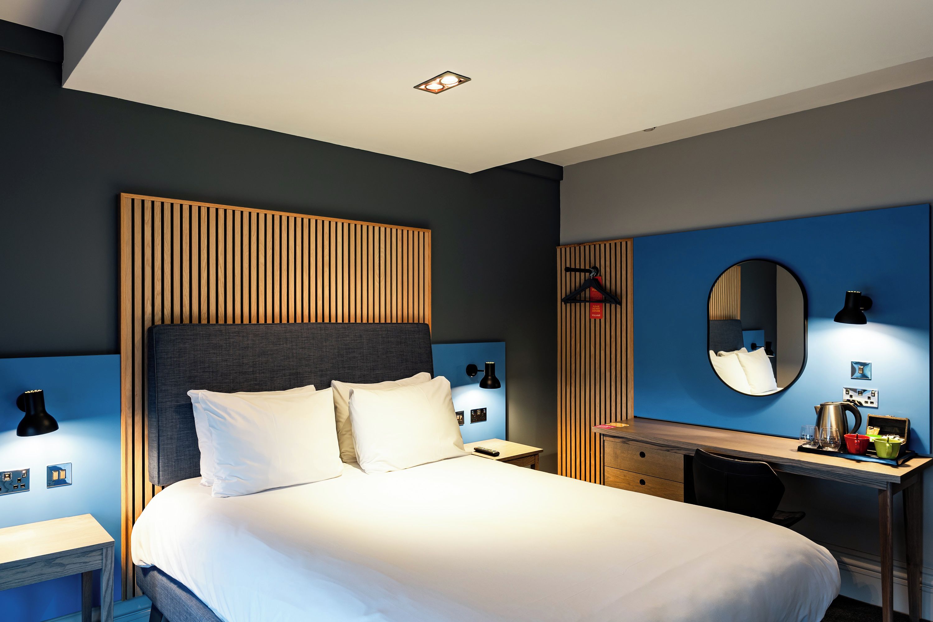 A modern hotel room with a large bed, a desk with a chair, a mirror, and a dresser.
