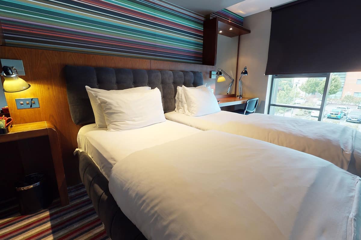 A hotel room with a double bed, a striped wall, a window with a view, and a nightstand with a lamp.