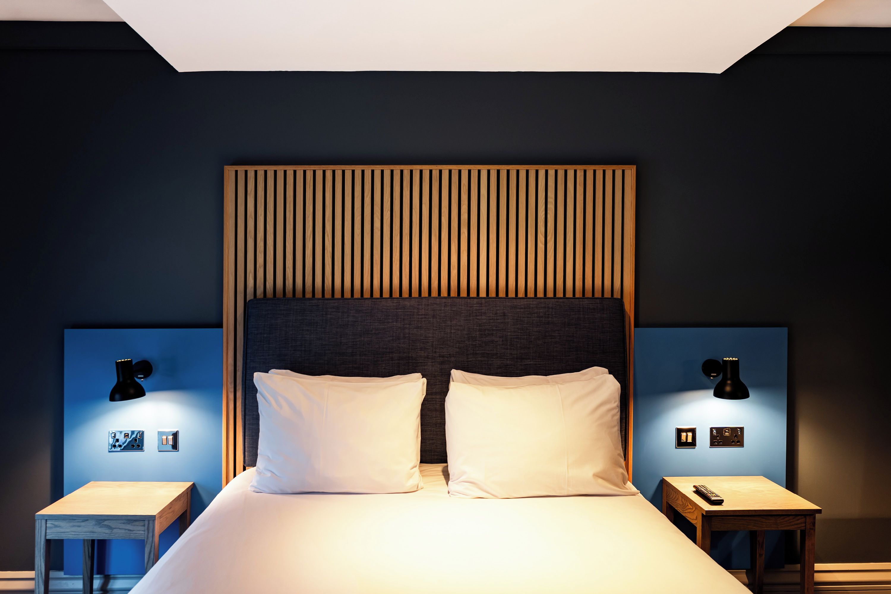 Quarto de hotel com cama, duas mesas de cabeceira, painel de cabeceira e luminárias fixadas na parede.