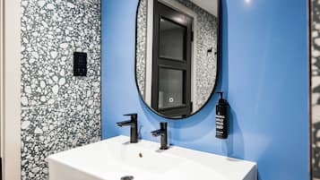 Un baño con un lavabo blanco, dos grifos negros, un espejo redondo y un dispensador de jabón en la pared.
