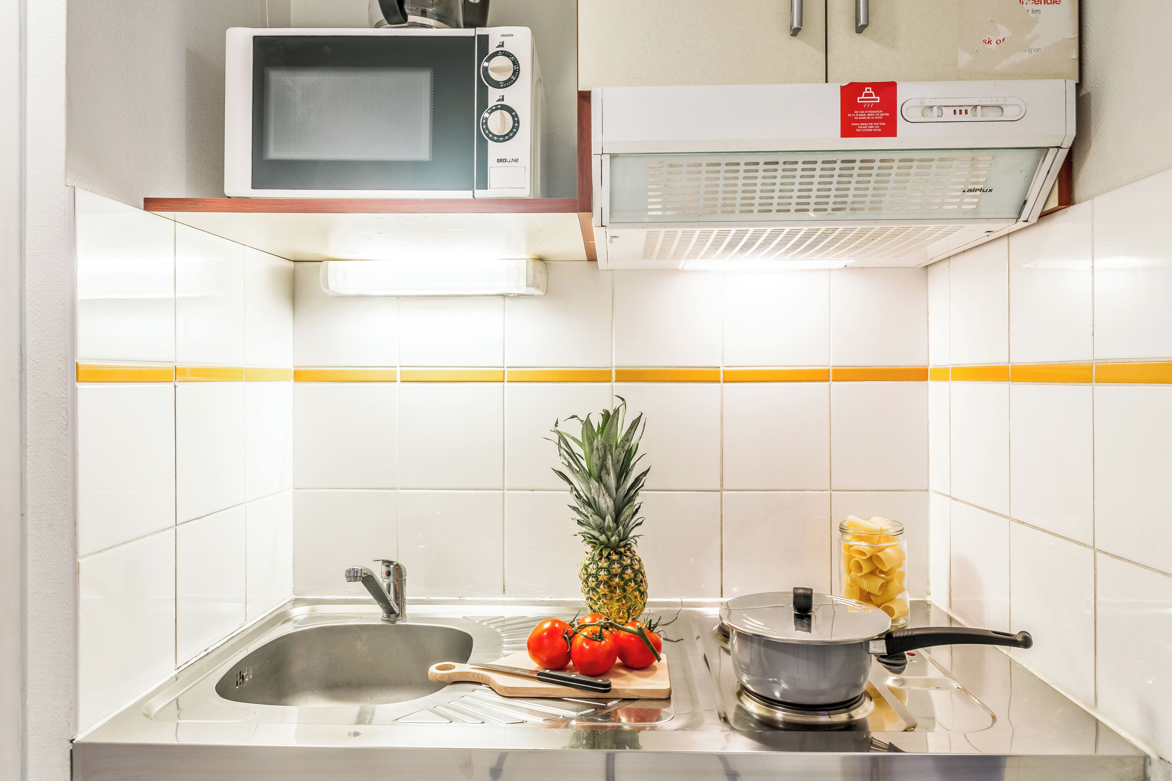 Een keuken met een magnetron, een fornuis, een spoelbak en een snijplank met tomaten en een ananas.
