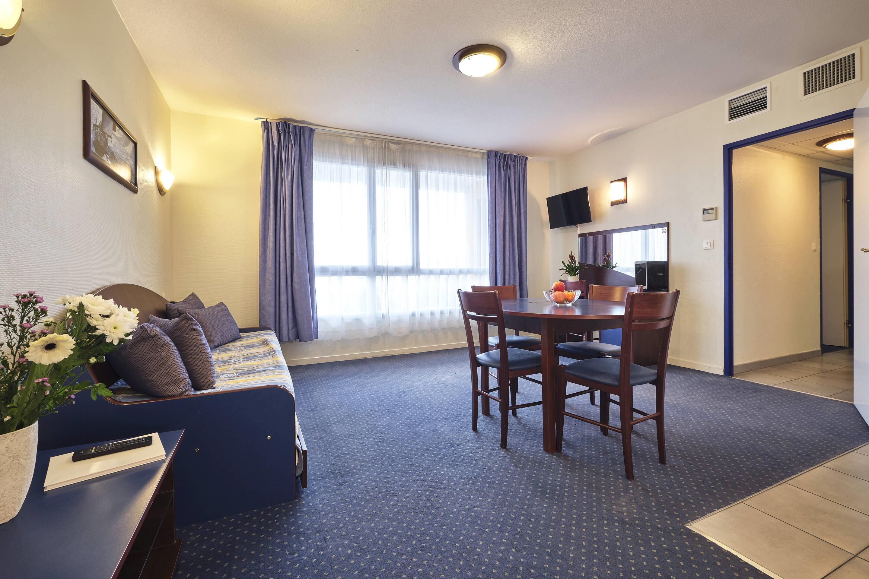 Quarto de hotel com sofá azul, mesa de jantar com cadeiras, televisão fixada na parede e uma janela grande com cortinas.