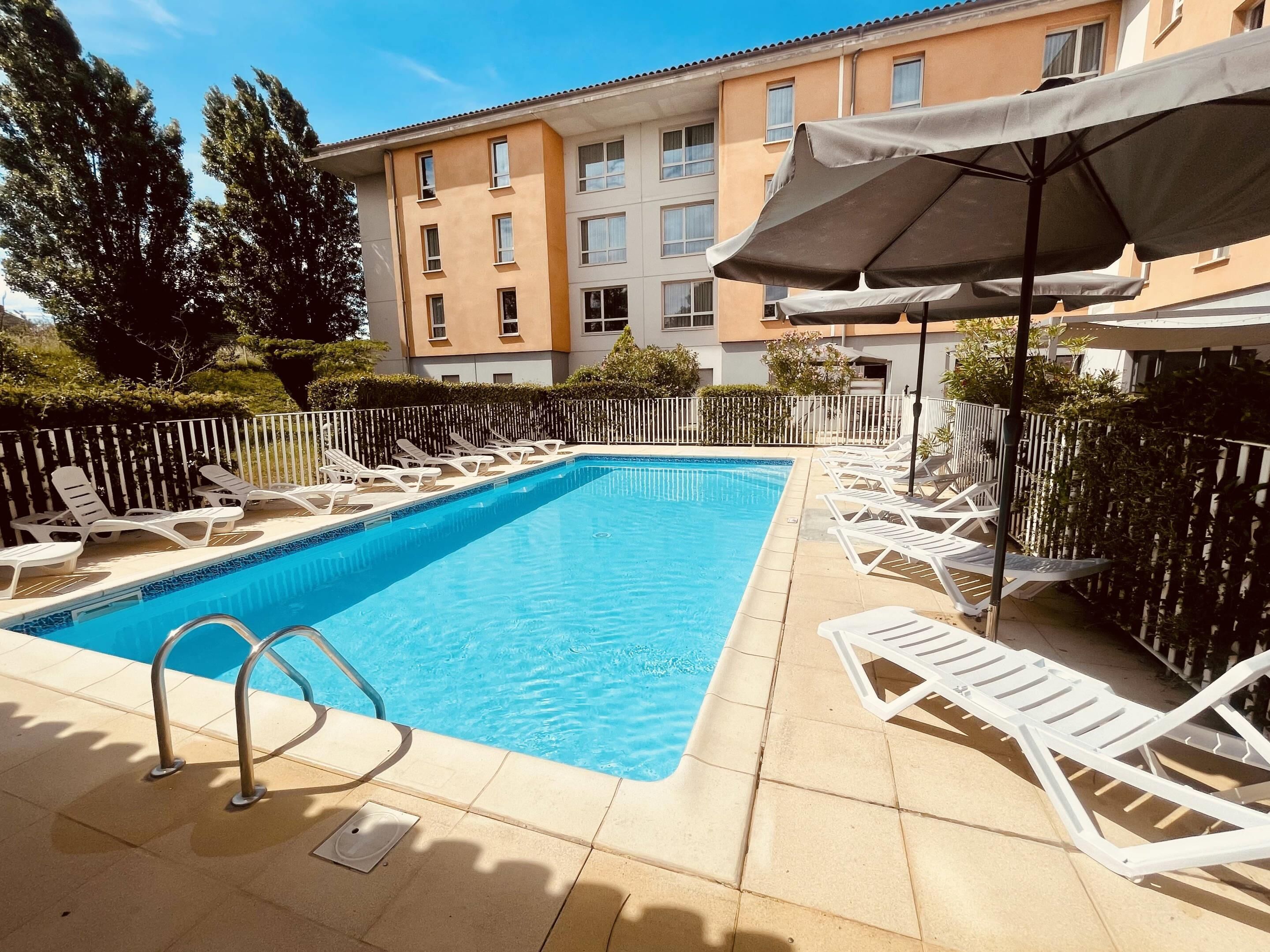 Una piscina rectangular con sillones blancos y sombrillas, rodeada por una cerca blanca y un edificio con múltiples ventanas.