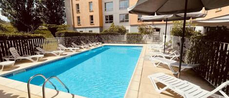 Una piscina rectangular con sillones blancos y sombrillas, rodeada por una cerca blanca y un edificio con múltiples ventanas.