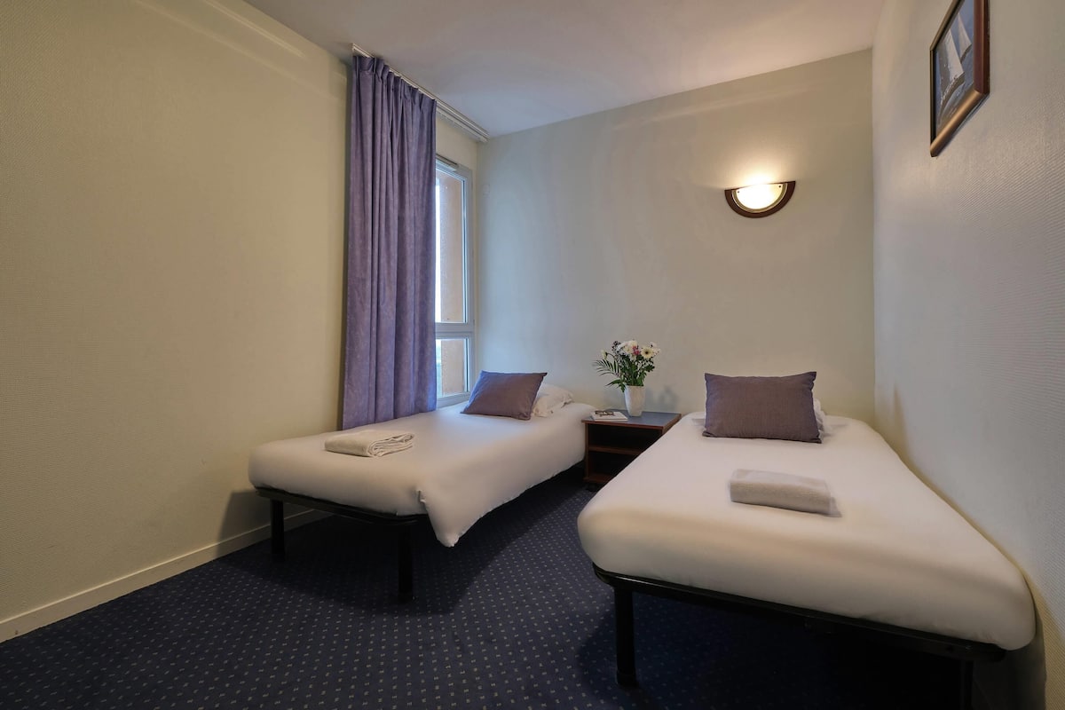 A hotel room with two single beds, a nightstand with a lamp, a window with curtains, and a wall-mounted light fixture.