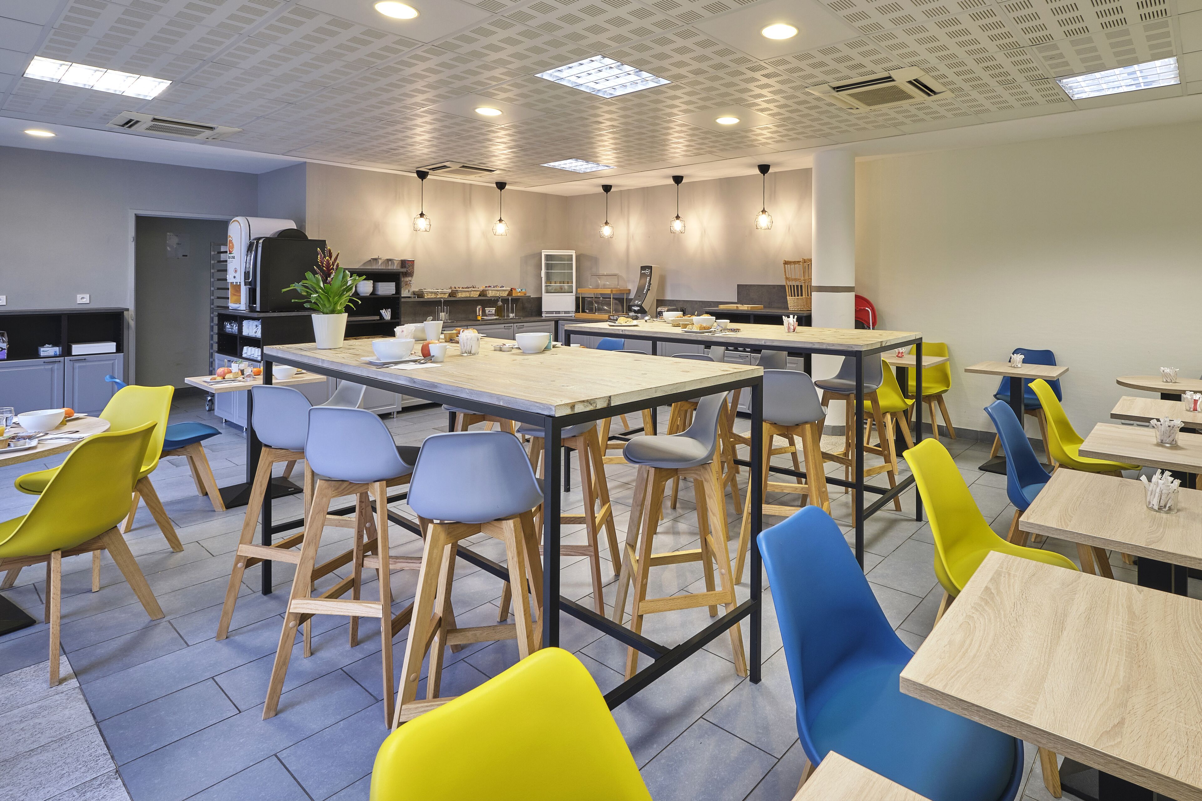 A modern dining area with multiple tables and colorful chairs, a kitchen in the background, and a ceiling with recessed lighting.