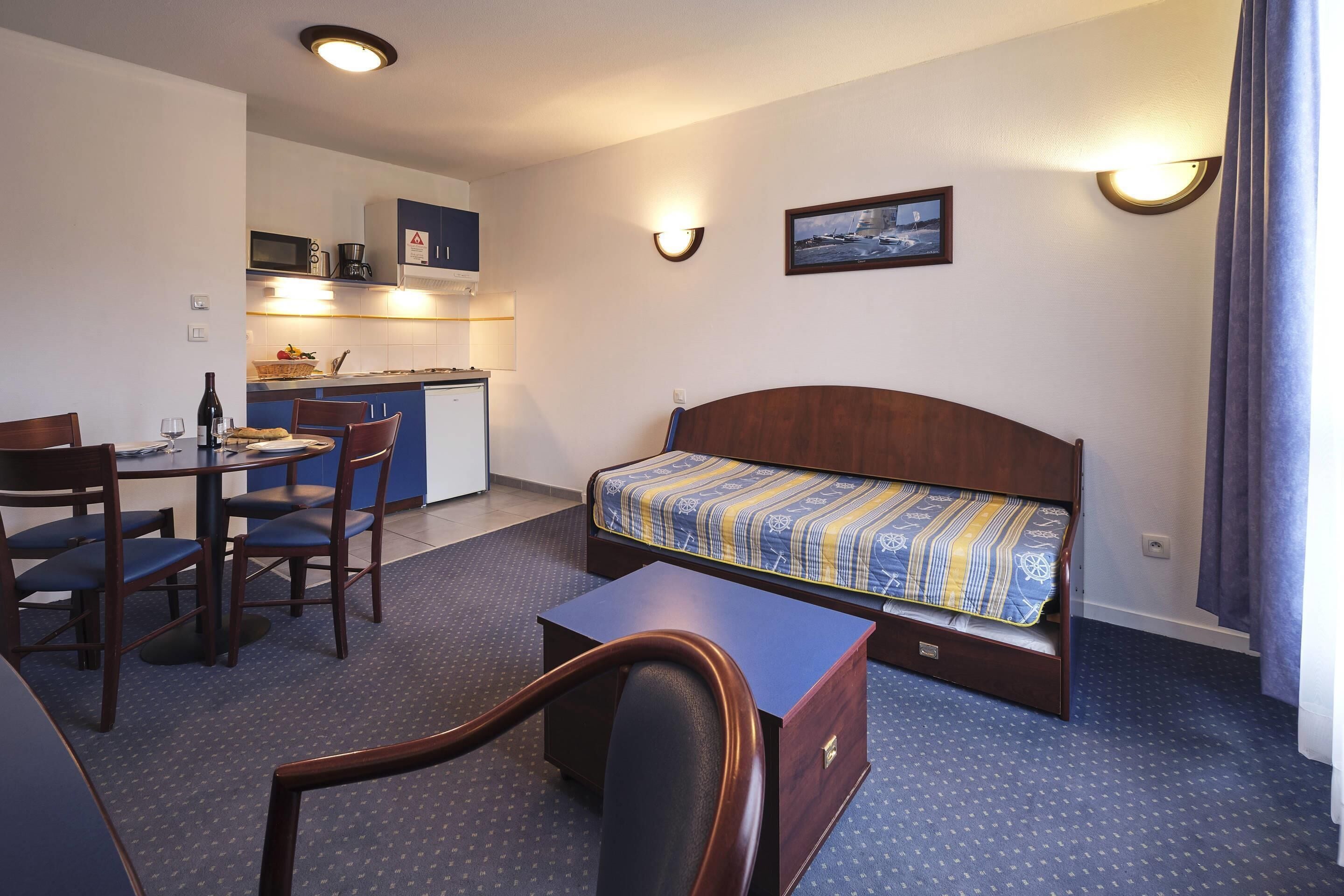 Quarto de hotel compacto com cama, pequena mesa de escritório, cadeira, mesa e cozinha compacta.