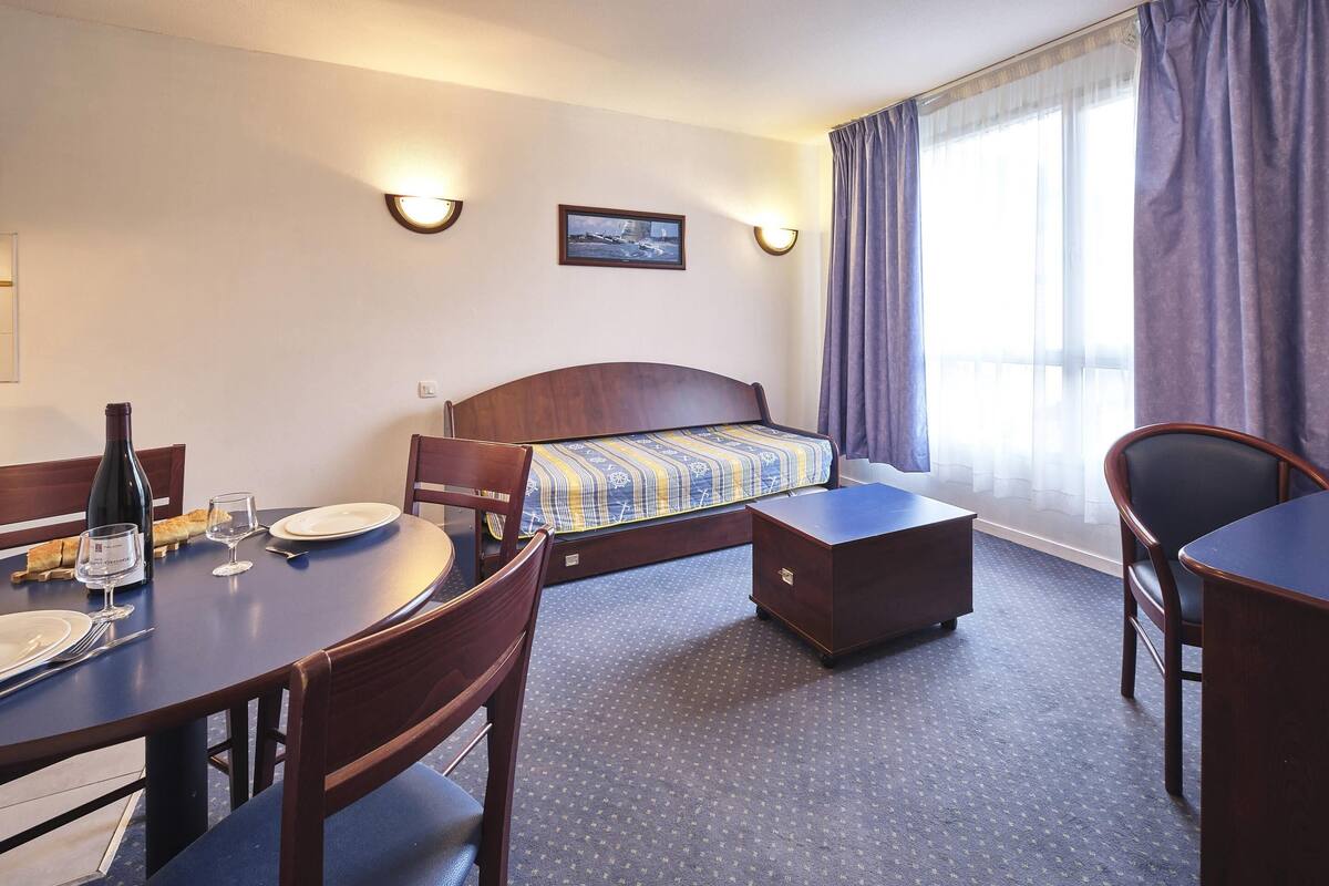 Een hotelkamer met een bed, een eettafel met stoelen, een bijzettafeltje met een fles en glazen, en een raam met gordijnen.