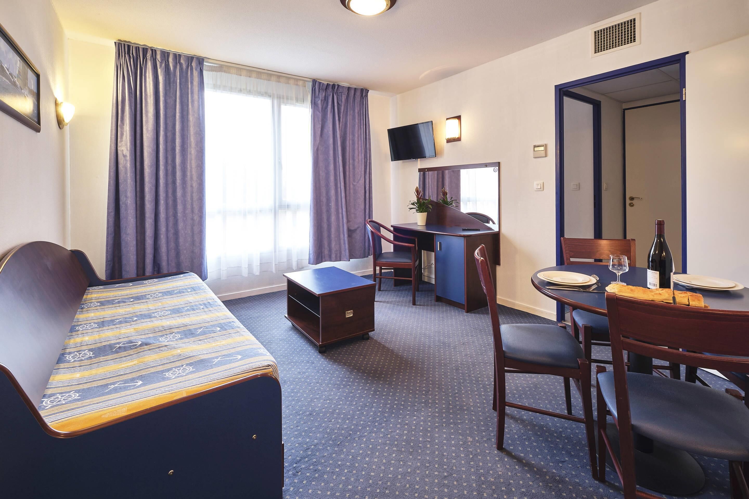 Quarto de hotel com cama, escrivaninha, cadeira, mesa de jantar com pratos e uma garrafa de vinho, e uma televisão na parede.