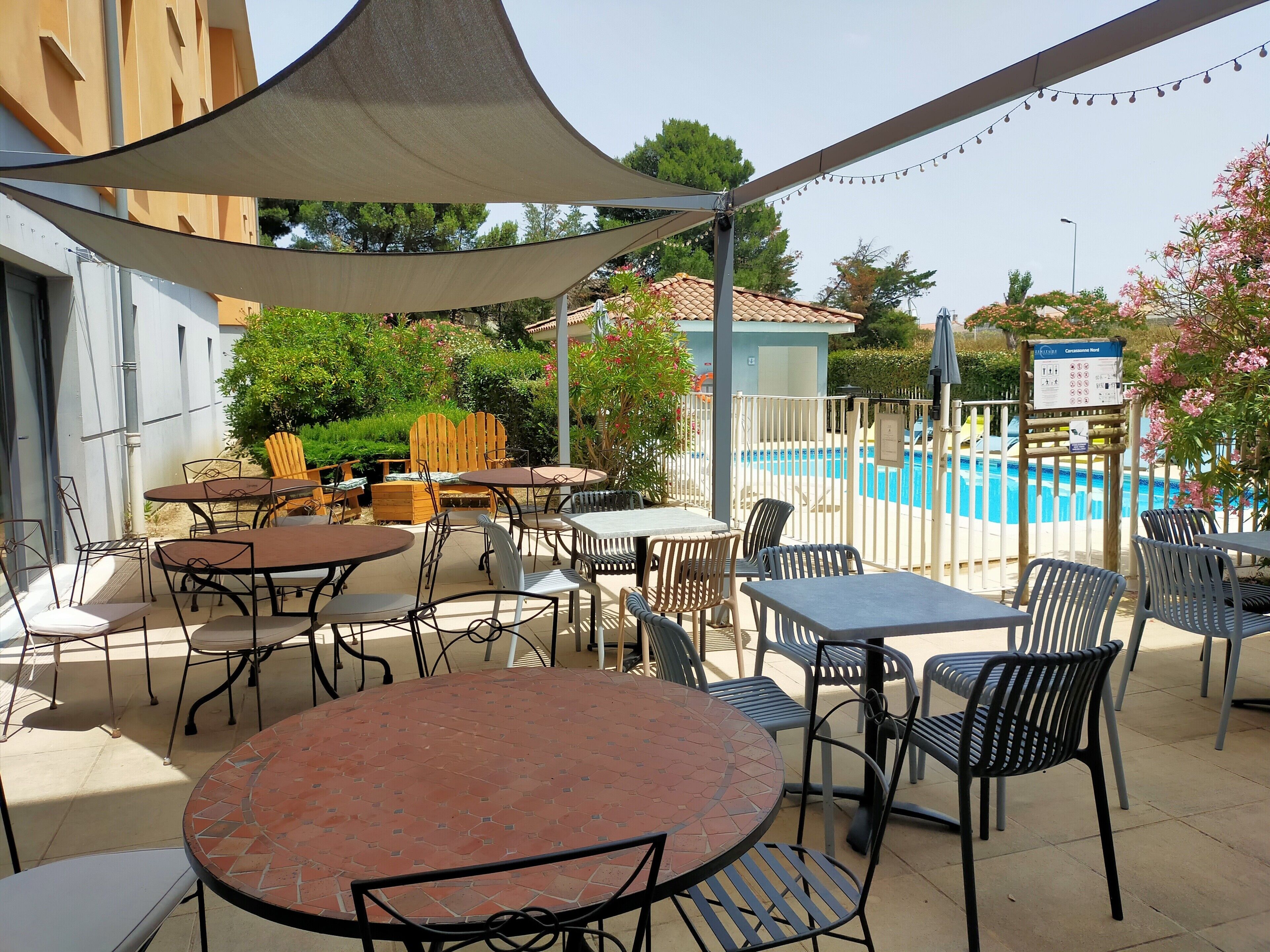 An outdoor patio area with multiple tables and chairs, a pool in the background, and a covered seating area.