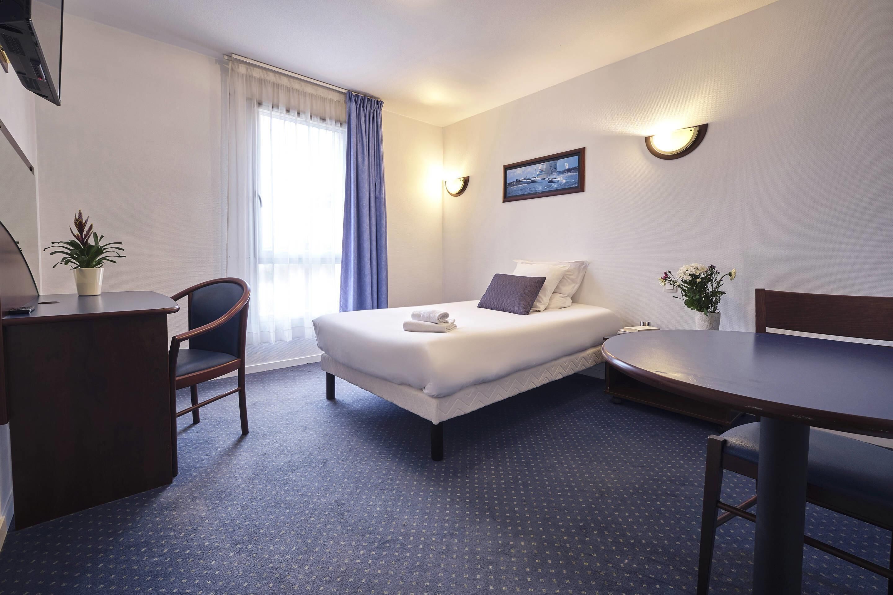 A hotel room with a bed, a desk, a chair, a table, and a potted plant.