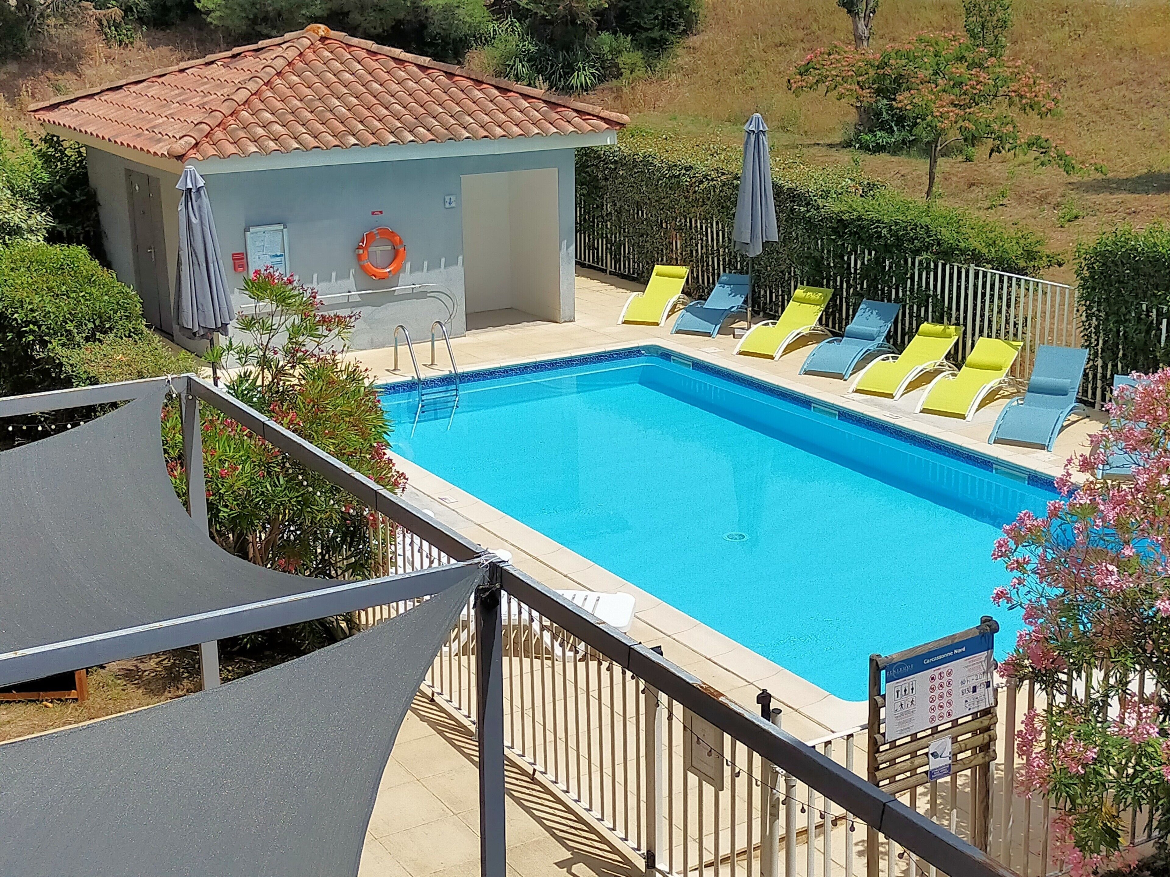 Ein Swimmingpool mit Fliesen umgeben, mit Liegestühlen und einem kleinen Gebäude mit einem gefliesen Dach.