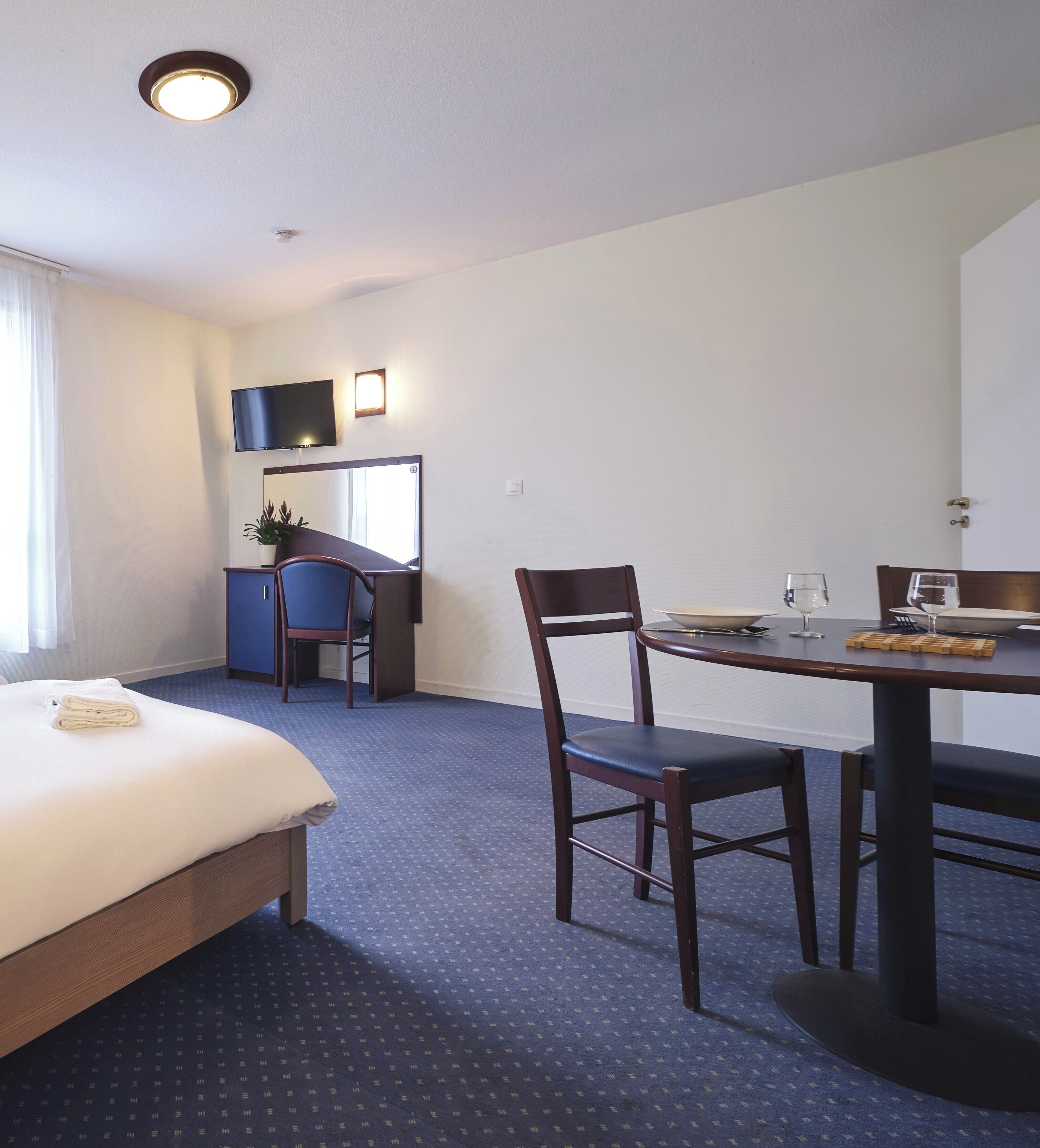 A hotel room with a bed, a desk, a chair, a dining table, and a TV.