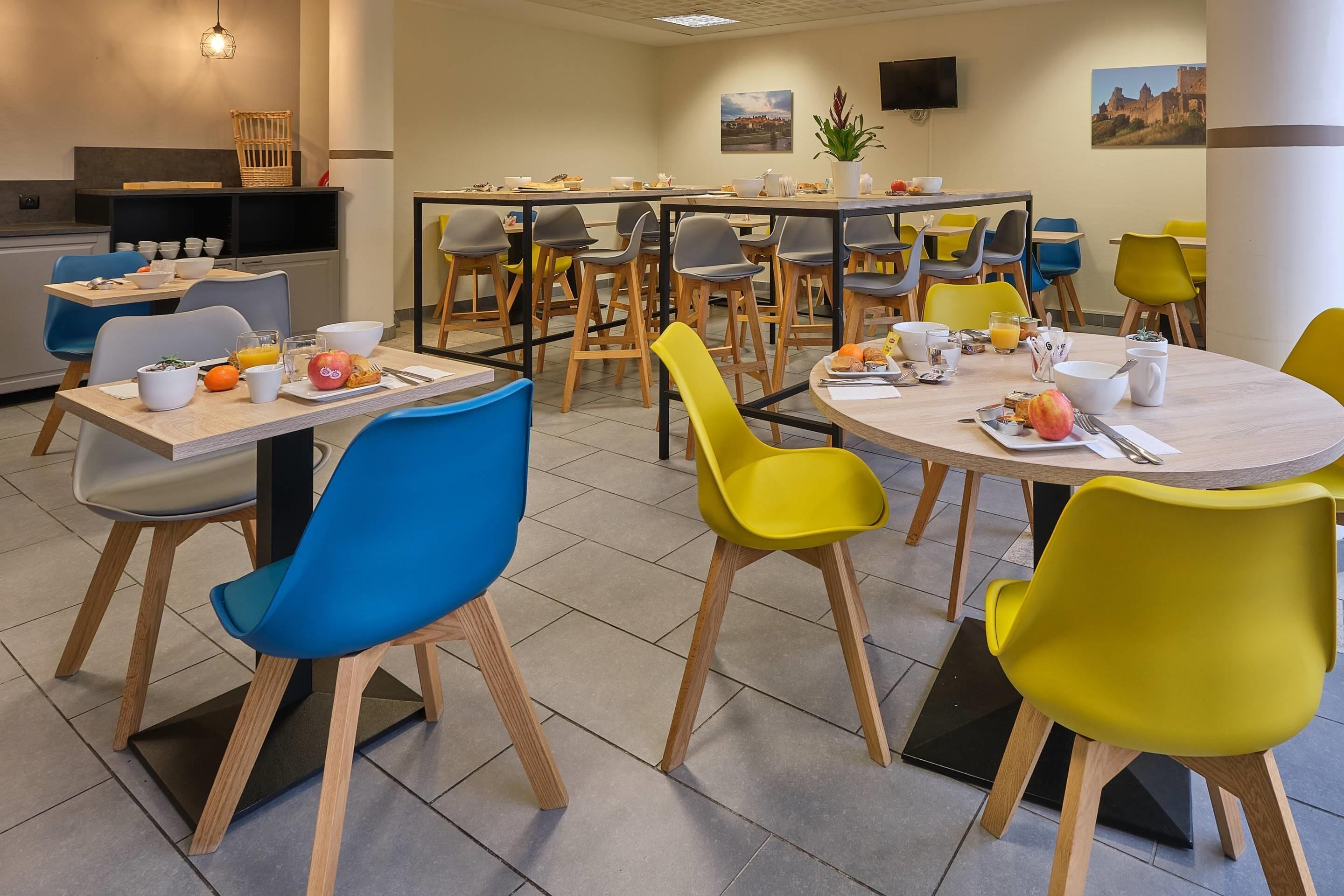 A modern dining area with multiple tables and chairs, equipped with basic amenities like cups, bowls, and fruits.