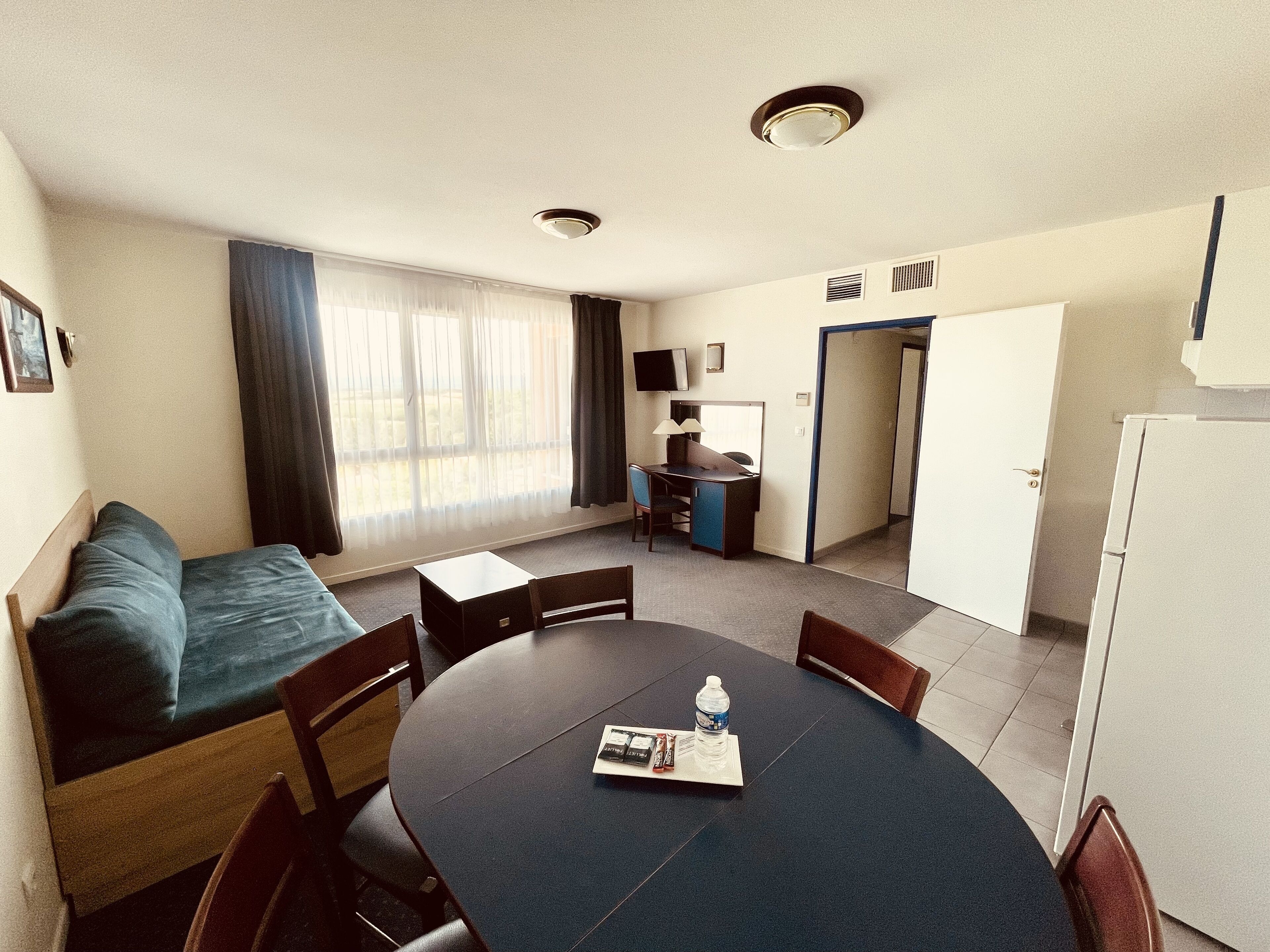 A compact hotel room with a bed, a dining table, chairs, a water bottle, and a television.