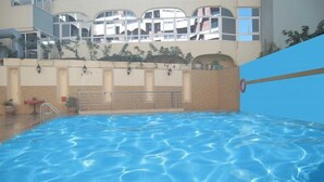 Ein Hallenbad mit klarem Wasser, einer Metallleiter und Blick auf ein Gebäude mit Balkonen und Pflanzen.