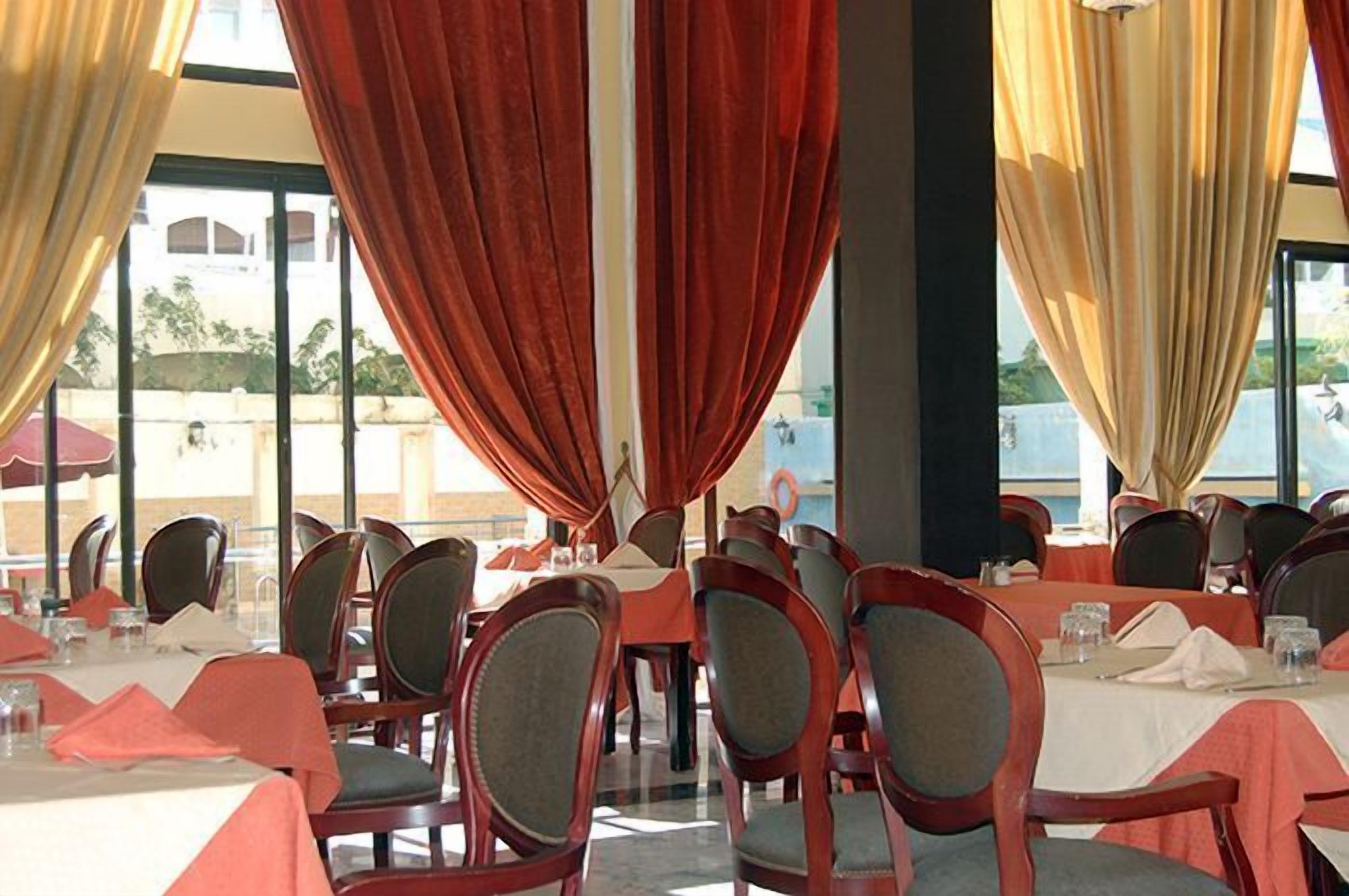 A restaurant with tables set for dining, red and beige curtains, and a view of the outside.