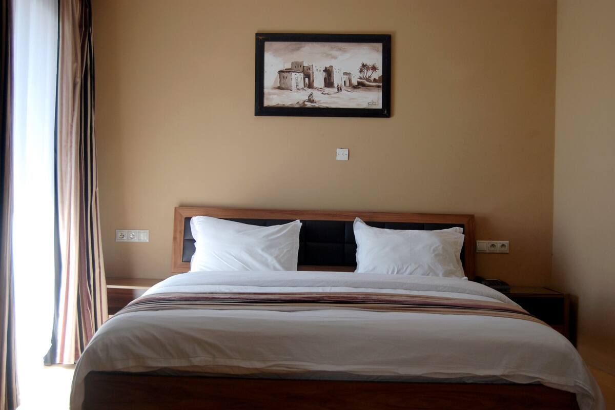 Ein Hotelzimmer mit Bett, Nachttisch, Wandbild und einem Fenster mit Vorhängen.