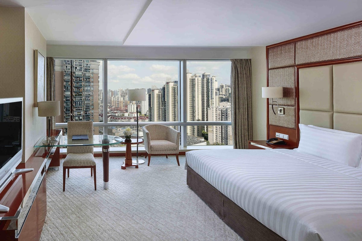 Habitación de hotel con una cama grande, un escritorio con televisor, una silla y vista a edificios altos.