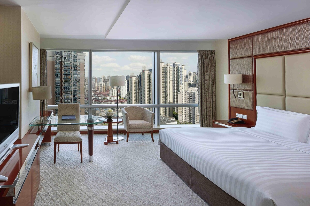 A hotel room with a large bed, a desk with a TV, a chair, and a view of tall buildings.
