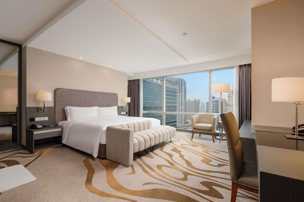 A modern hotel room with a large bed, a desk, a chair, and a view of the cityscape.