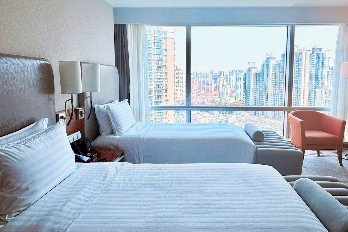 Habitación de hotel con dos camas, un sofá, una silla y un ventanal amplio con vistas a la ciudad.