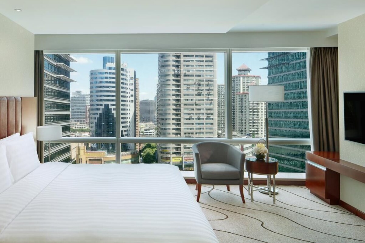 Habitación de hotel con una cama grande, una silla, una mesita y un televisor. La habitación tiene ventanas amplias con vista a la ciudad.