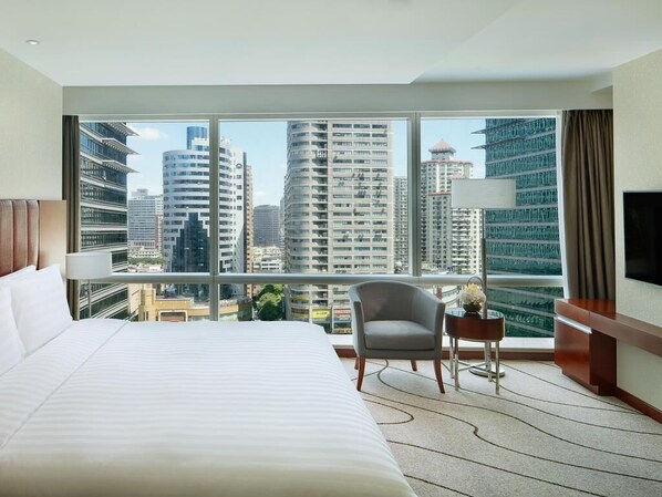 Une chambre d’hôtel avec un grand lit, une chaise, une petite table et un téléviseur. La chambre dispose de grandes fenêtres donnant sur la ville.