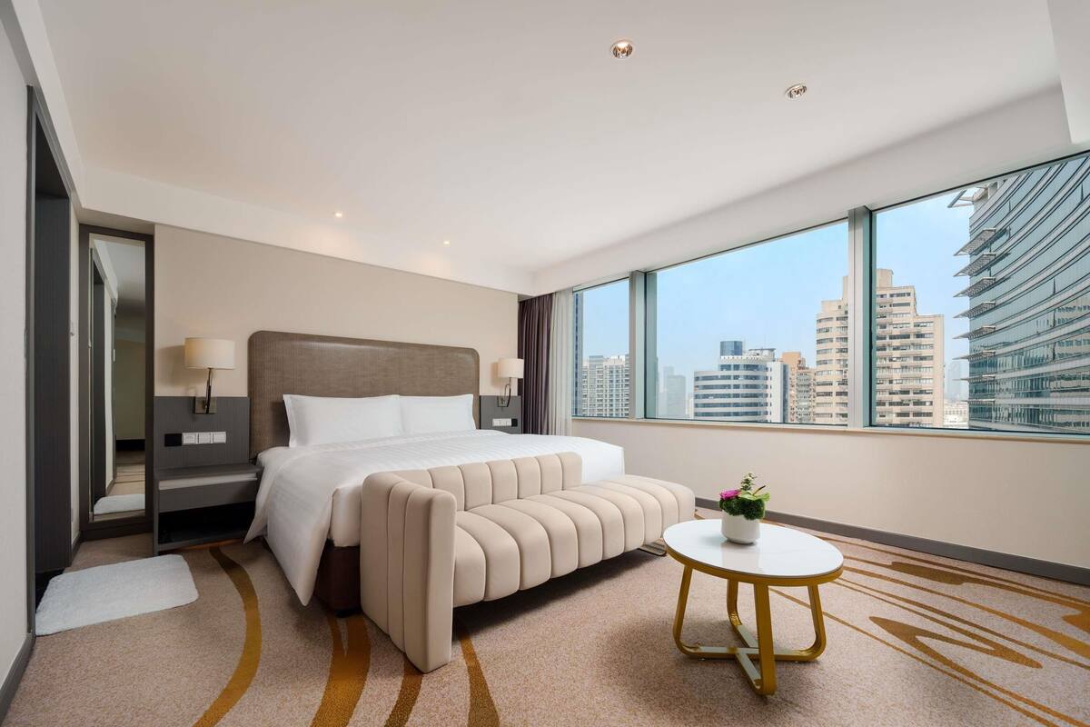 Habitación de hotel moderna con una cama grande, una mesita de noche con lámparas, una pequeña mesa redonda y vistas a la ciudad a través de amplios ventanales.