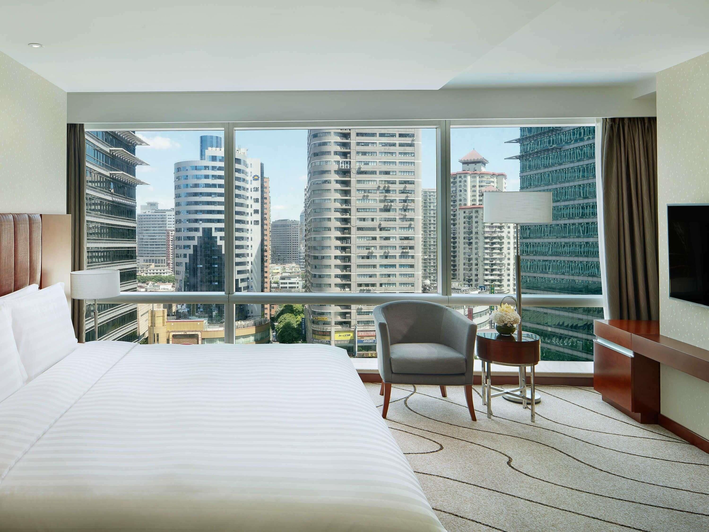 A hotel room with a large bed, a chair, a small table, a TV, and a view of tall buildings.