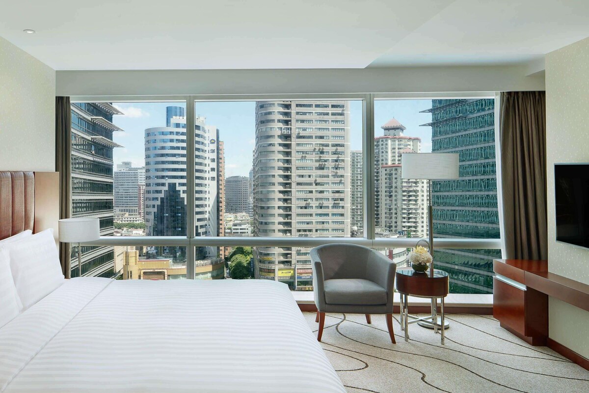 Quarto de hotel com cama grande, uma cadeira, mesa pequena, TV e vista para edifícios altos.