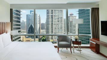 A hotel room with a large bed, a chair, a small table, a TV, and a view of tall buildings.