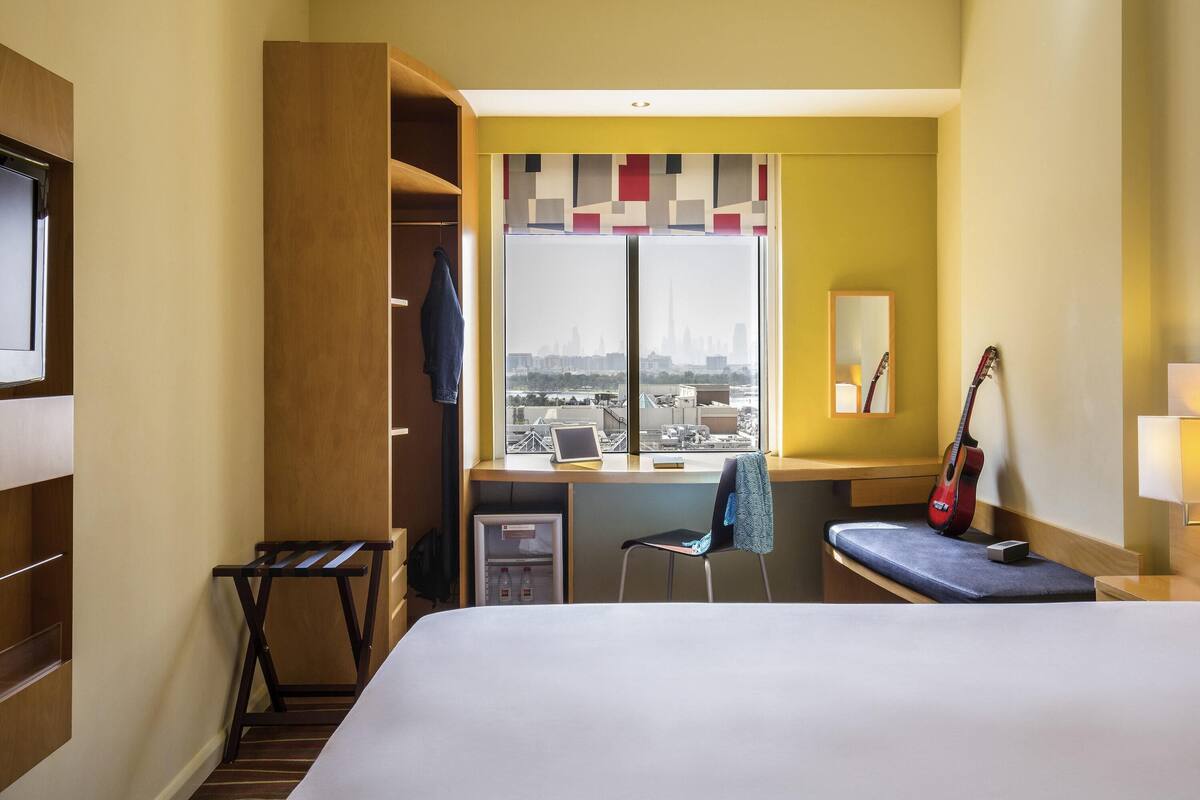 A hotel room with a bed, a desk, a chair, a guitar, a wardrobe, and a window with a city view.