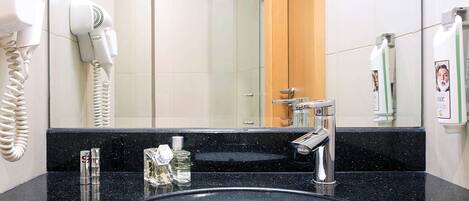A modern bathroom with a black granite countertop, a white sink, a handheld hair dryer, and a mirror reflecting a wooden door and a glass shower enclosure.