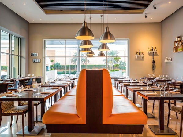 A restaurant with multiple tables, a central orange bench, pendant lighting, and large windows with a view of palm trees. - ibis Deira Creekside Dubai (Dubai)
