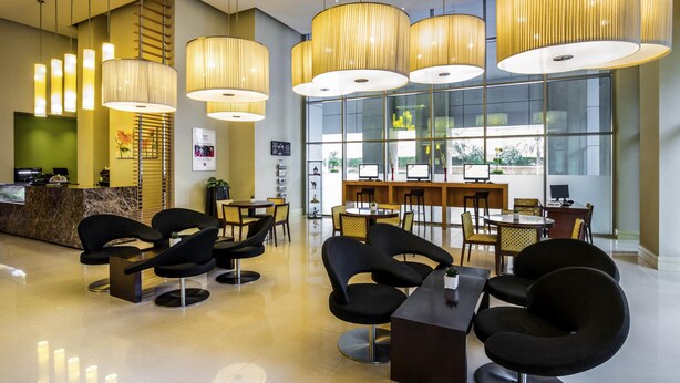 Um saguão de hotel moderno com balcão de recepção, cadeiras pretas, uma mesa comprida e luminárias pendentes grandes.