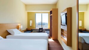 Quarto de hotel com duas camas, uma escrivaninha, uma cadeira, uma televisão e uma janela grande com vista para a cidade.