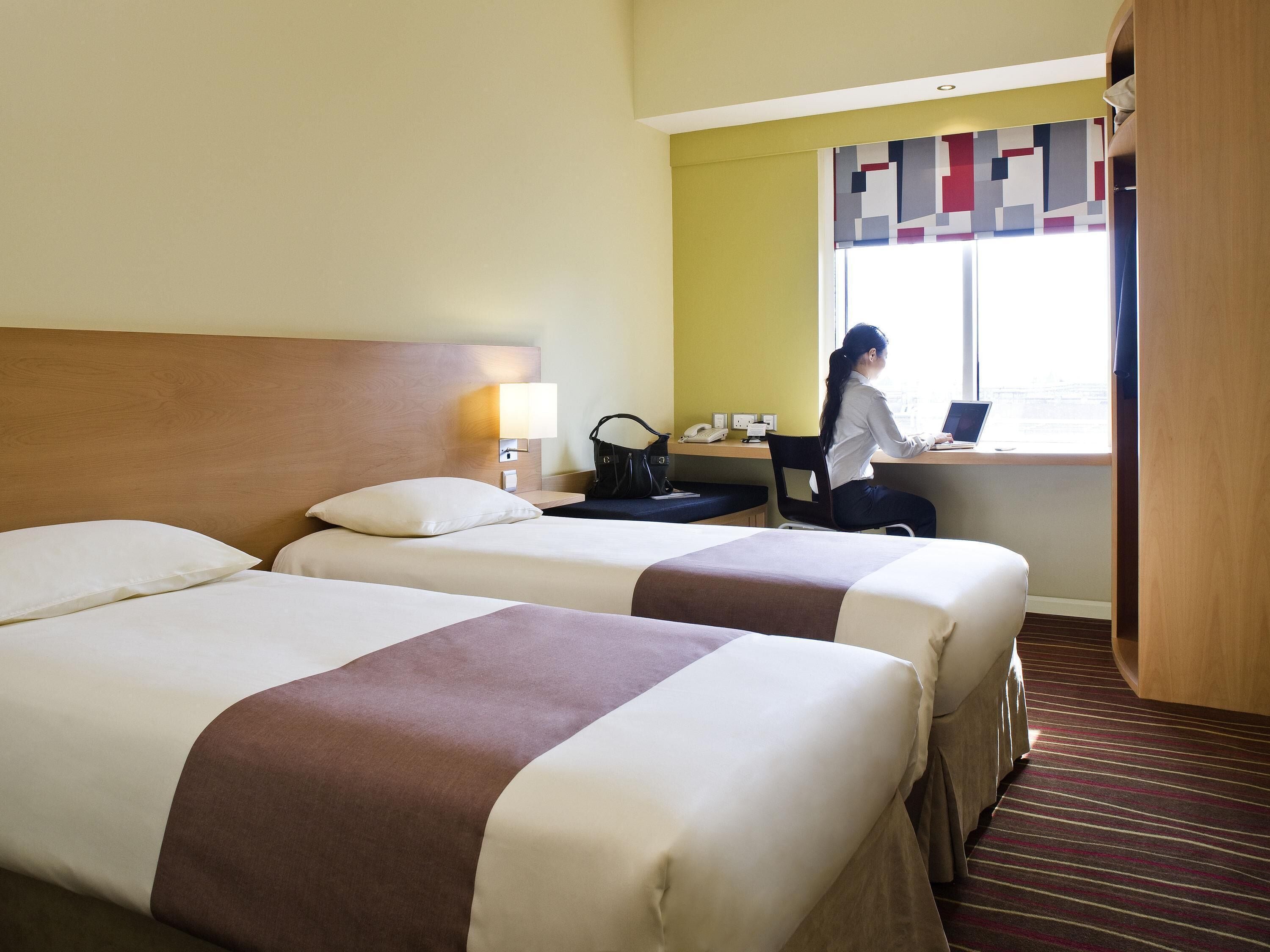 Une chambre d’hôtel avec deux lits, un bureau avec un ordinateur portable, un téléphone et une fenêtre avec des rideaux.