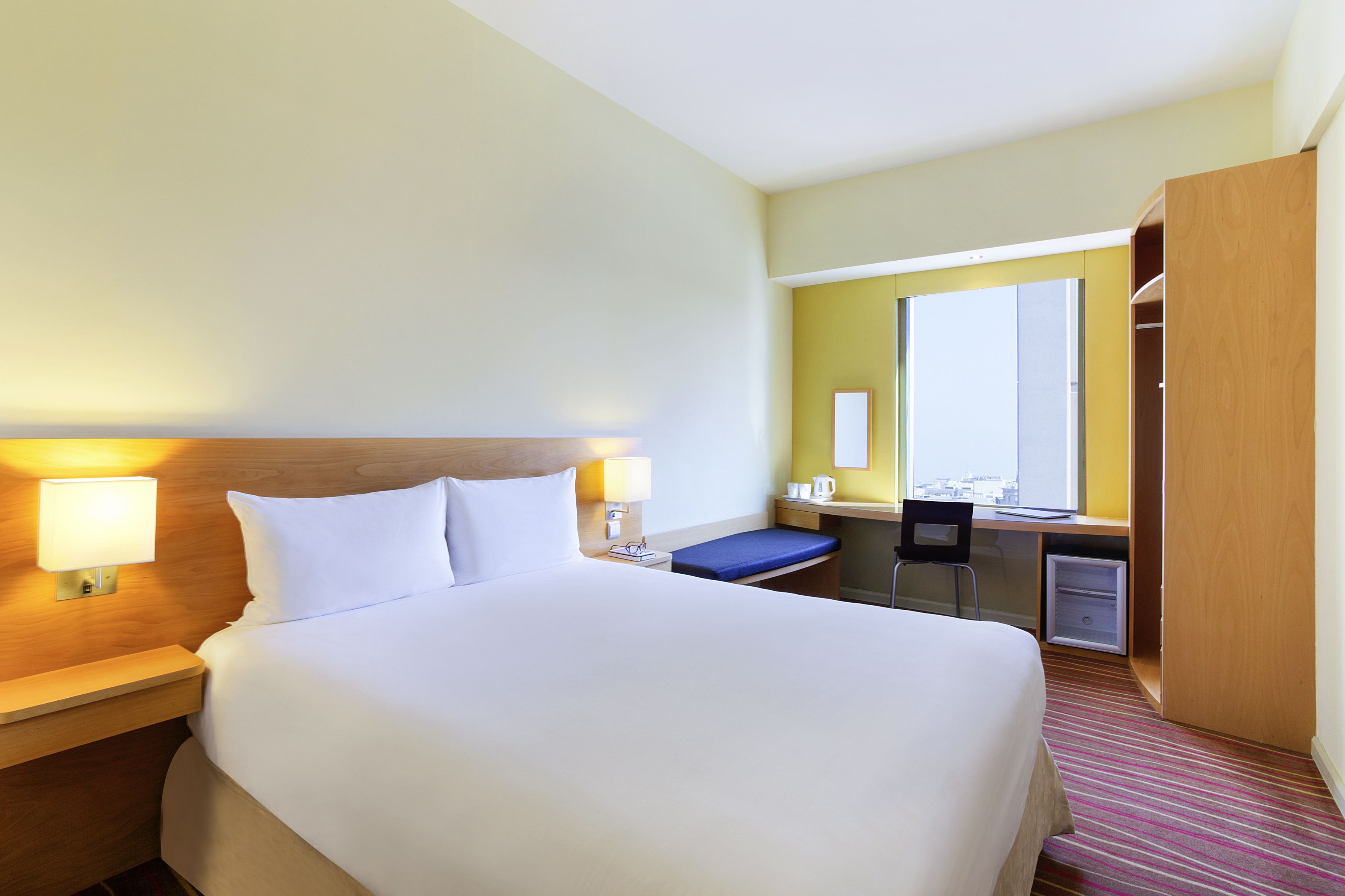 A hotel room with a large bed, bedside lamps, a desk with a chair, a mirror, and a wardrobe.