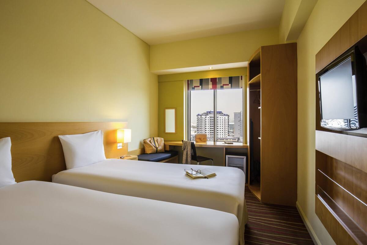 Hotellihuone, jossa on kaksi sänkyä, työpöytä, tuoli, vaatekaappi ja suuri ikkuna.